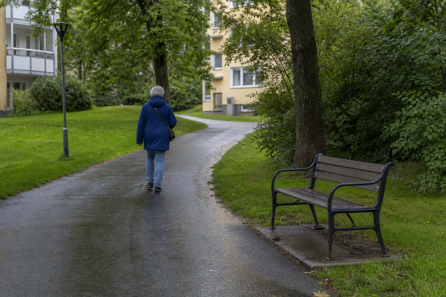 En äldre person med en liten axelväska promenerar på en våt gångväg förbi en tom parkbänk.